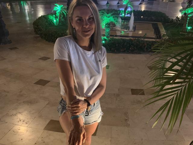 BelleLisaG, em pé com as mãos unidas na frente, vestindo uma camiseta branca e shorts jeans curtos, olhando diretamente para a câmera, ligeiramente inclinada para frente, com pouca luz, mostrando plantas iluminadas em verde..