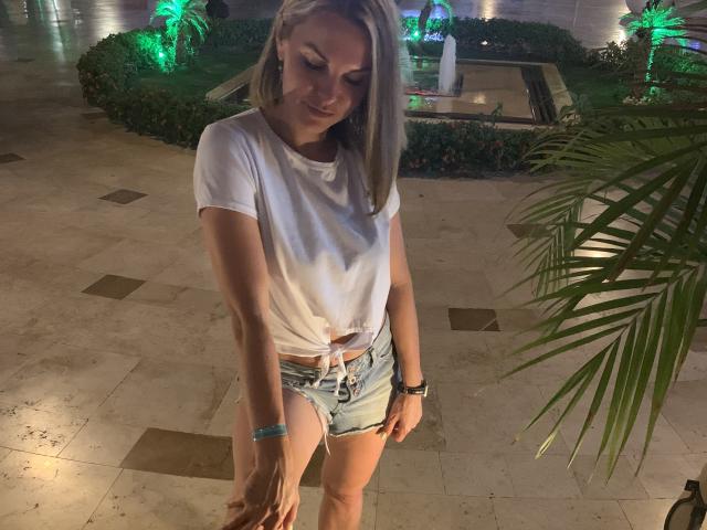 BelleLisaG, standing indoors at night, wearing a white t-shirt tied at the front and denim shorts, looking down with a slight smile, green-lit garden fountain in the background. Sexual tension evident.