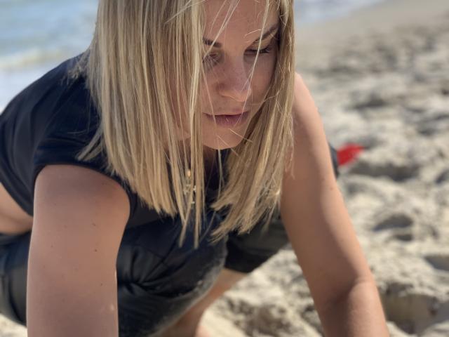 BelleLisaG, avec ses cheveux blonds mi-longs, accroupie sur une plage de sable, portant un crop top noir, avec du sable dans les cheveux, regard intense, reflets de la lumière du soleil sur les mèches de cheveux, fond avec une plage..