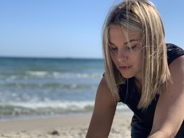 BelleLisaG, avec ses courts cheveux blonds, se penche en avant sur une plage, vêtue d'un haut noir. Ses yeux sont baissés, créant une atmosphère sensuelle et tendue. Des vagues de l'océan en arrière-plan. Une lumière du soleil vive..