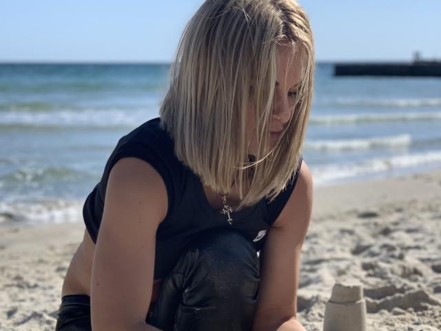 Blonde BelleLisaG, crouched on sandy beach, wearing black wet shirt, building sandcastle, ocean waves in background, sunlight reflecting off hair, sexual tension, beach setting, summer vibes, sand on skin.