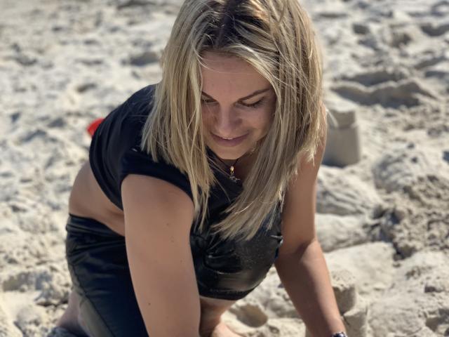 BelleLisaG, avec des cheveux blonds, penchée en avant sur une plage de sable, portant un crop top noir et un short, éclairée par le soleil, avec une ombre sur le visage, expression sérieuse, reflets lumineux dans les cheveux, lumière du soleil vive..