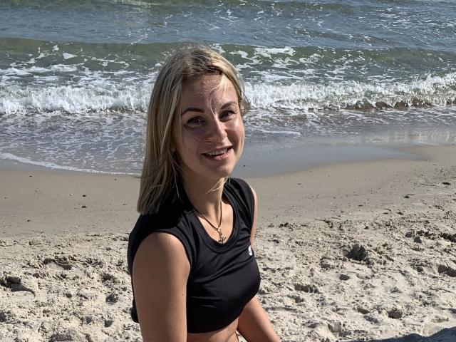 Blonde BelleLisaG, smiling at the camera, stands on a sandy beach with waves crashing behind her. She wears a black crop top, sunlight glistening on her skin, beach sand on her shoulders.