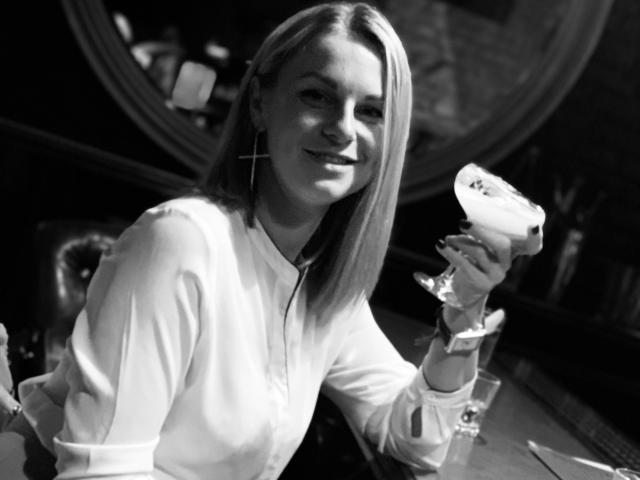 Black-and-white photo of BelleLisaG, smiling with a glass of white liquid in her right hand, wearing a light-colored, long-sleeve shirt, dimly lit background with circular shapes. Fucking hot.
