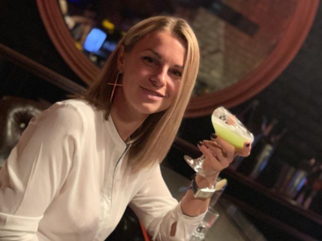 BelleLisaG, une femme blonde aux cheveux lisses, portant un haut blanc à manches longues, tenant un cocktail jaune dans un verre, souriante. Légèrement sombre, avec un bar éclairé, un miroir rond et un arrière-plan flou, créant une ambiance séduisante..