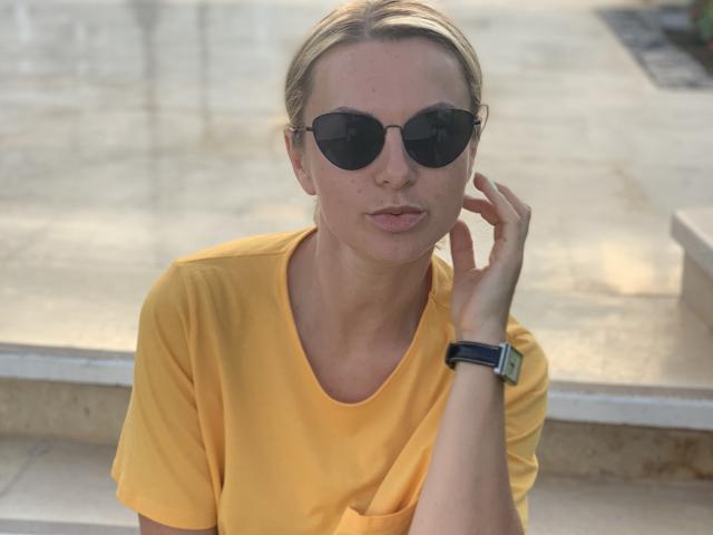 Photograph of BelleLisaG, wearing a yellow t-shirt, black sunglasses, and a black smartwatch. She's touching her face with her left hand, exuding a casual, slightly sultry vibe. Bright sunlight.