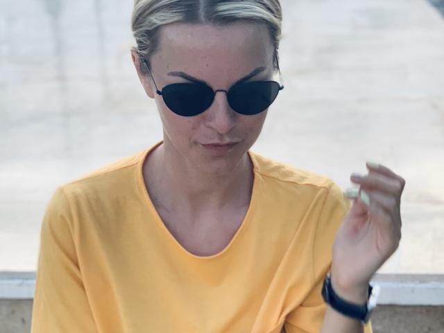 Photograph of BelleLisaG, blonde, wearing black sunglasses, yellow shirt, holding a cigarette in right hand, wristwatch on left wrist, focused gaze, bright light background, casual, slightly sexy vibe.