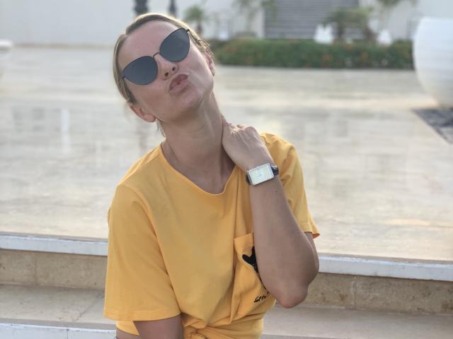 BelleLisaG está encostada em uma janela, vestindo uma camiseta amarela e óculos de sol escuros, com a cabeça inclinada para trás e a mão apoiada. seu pescoço, transmitindo uma aura de confiança e sensualidade. A luz do sol realça sua beleza..