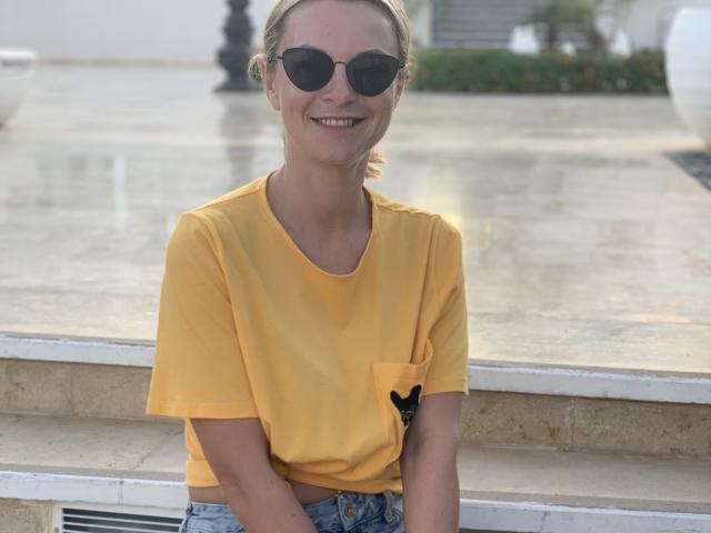 BelleLisaG está sentada ao ar livre, sorrindo, vestindo uma camiseta amarela com um bolso preto e óculos de sol redondos. Ela está apoiada em uma beirada de pedra, com um fundo desfocado. Suas calças jeans são visíveis. Muito fofo.