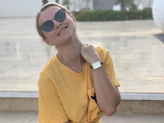 BelleLisaG se apoya contra una ventana de vidrio, sonriendo con la cabeza inclinada hacia atrás. Ella lleva una camisa amarilla, gafas de sol y un reloj de plata. Su mano descansa sobre su cuello, irradiando una sensualidad casual..