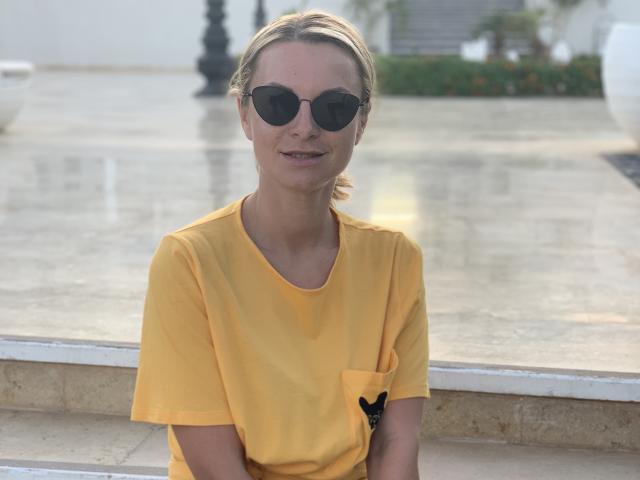 Photographie de BelleLisaG, une femme à la peau claire et aux cheveux bruns attachés en queue de cheval, portant des lunettes de soleil rondes et un vêtement jaune. T-shirt avec une poche noire, porté à l'extérieur par un jour ensoleillé..