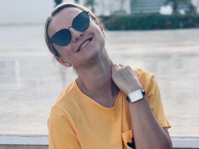 BelleLisaG, portant un t-shirt orange et des lunettes de soleil noires, sourit, la tête penchée en arrière, une main sur le cou. Elle porte une montre argentée à son poignet, avec la lumière du soleil en arrière-plan. Extrêmement sexy.