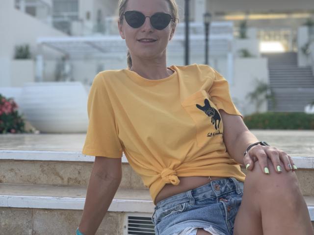 BelleLisaG, sentada em um banco de pedra, vestindo uma camisa amarela de amarração, shorts jeans e óculos de sol, com unhas de um tom de verde neon. polimento, transmitindo uma vibe relaxada e sensual, ao ar livre, ensolarado..