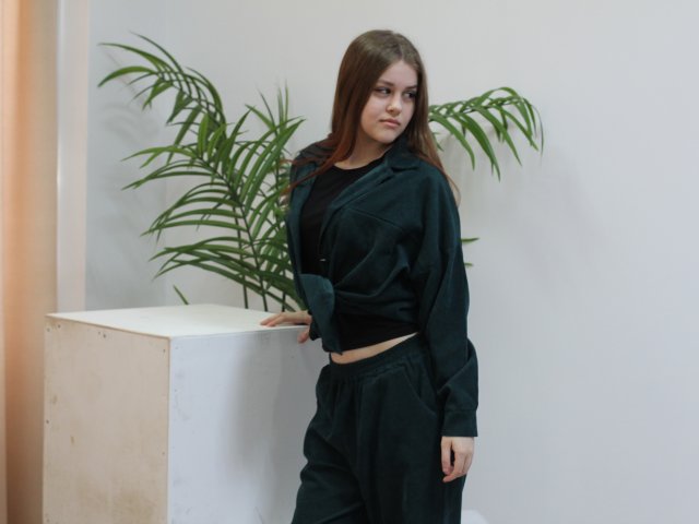 LilaFraser, met lang bruin haar, staat tegen een witte muur, leunend op een witte sokkel. Ze draagt een zwarte outfit en ze ziet er serieus uit, met een groene plant achter haar. Heel aantrekkelijk.