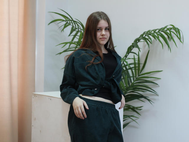 Photographie de LilaFraser, avec de longs cheveux bruns, debout devant un mur blanc et une plante verte en pot. Portant une chemise et un pantalon noirs, une ceinture blanche, dégageant une tension sexuelle, les mains sur les hanches..