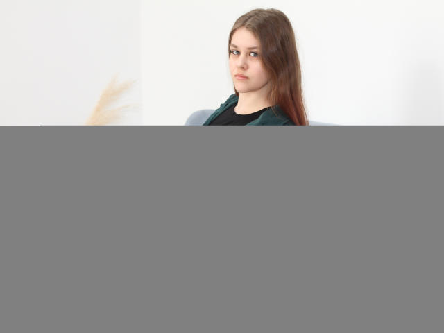 Lila Fraser, met lang bruin haar, gekleed in een zwarte outfit, zittend op een lichtblauwe, gestoffeerde bank, leunend achterover met een neutrale uitdrukking, pluizige beige pampa gras op de achtergrond..