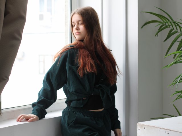 LilaFraser staat bij een helder raam, gekleed in een zwarte outfit met lange mouwen en een broek met hoge taille. Haar lange, bruine haar valt over haar schouder, haar ogen zijn gesloten, en zacht licht verlicht haar gezicht. Potplant op de achtergrond.