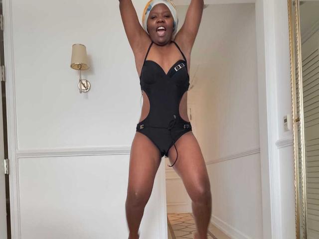 AfricanaKisses, dark-skinned woman with muscular build, wearing a black one-piece swimsuit with cutouts, arms raised, smiling with mouth open, white beanie, standing in a white-walled room. Fierce, confident stance.