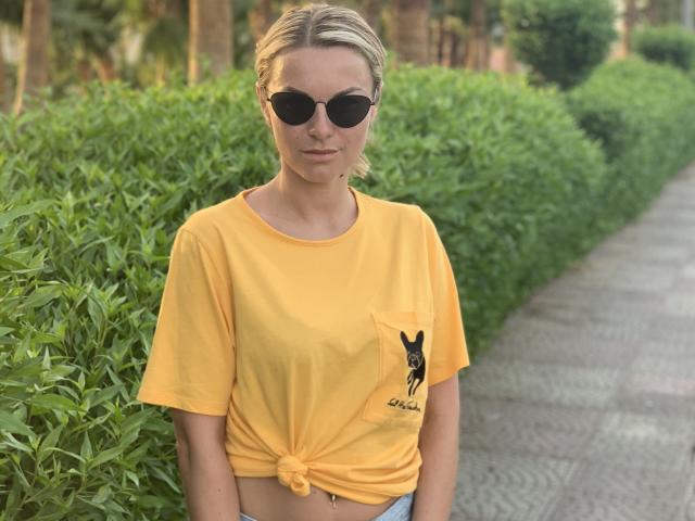 BelleLisaG avec des tresses blondes, portant un t-shirt jaune noué à la taille, des lunettes de soleil noires, debout devant un fond vert. buissons. Expression concentrée, légèrement confiante, cadre extérieur décontracté..