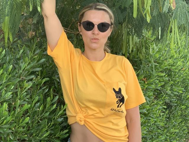 BelleLisaG, uma mulher esguia de pele clara e cabelos loiros, veste uma camiseta amarela com uma estampa preta, amarrada na cintura, e óculos de sol escuros. Ela levanta o braço, cercada por uma vegetação verde exuberante. Que demais..