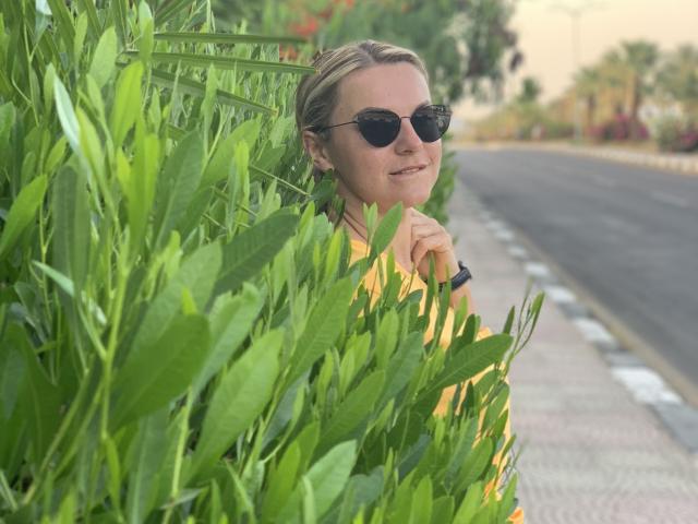 BelleLisaG escondida atrás de uma folhagem verde densa, usando óculos de sol pretos, com um sorriso discreto. A estrada e o fundo desfocado são visíveis. A mão dela está próxima ao queixo, transmitindo uma vibe casual e sensual..