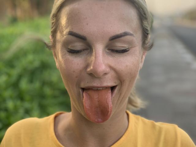 Close-up of BelleLisaG with sweat-covered face, eyes closed, tongue sticking out. She wears a yellow shirt, has blonde hair, and a teasing expression. Blurred green and gray background.