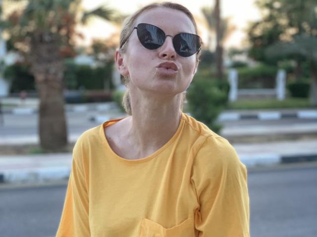 BelleLisaG, photo en gros plan, portant une chemise jaune, des lunettes de soleil noires, les lèvres pincées, en extérieur, avec des palmiers flous en arrière-plan. et arrière-plan de rue, lumière naturelle, style décontracté, léger angle, expression confiante.