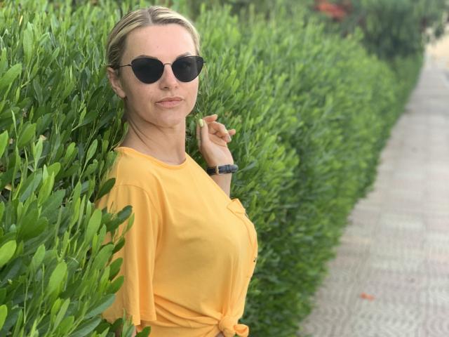 BelleLisaG, standing against lush green bushes, wearing a yellow top, black sunglasses, and a black wristwatch, smiling slightly with a subtle hand gesture. Bright, sunny outdoor setting.