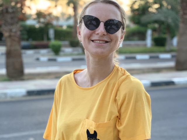 BelleLisaG indossa una camicia giallo brillante con un ricamo a forma di gatto nero sulla tasca, sorride con occhiali da sole scuri, ed è in piedi. all'aperto.
lo sfondo include alberi sfocati e asfalto. Atmosfera estiva, molto sensuale..