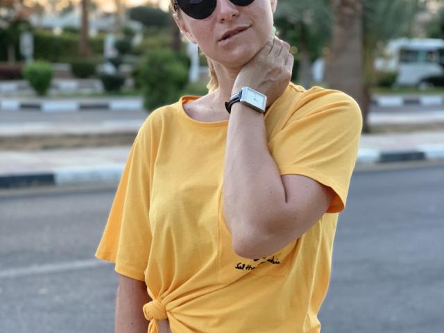 BelleLisaG wearing a bright yellow t-shirt tied at the waist, black sunglasses, and a white wristwatch, stands with her hand on her neck, exuding confident, sexy vibes. Background blurred.