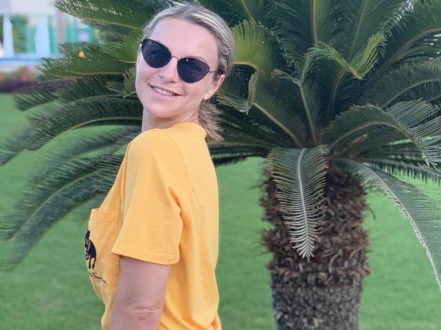 BelleLisaG com cabelo loiro preso em um rabo de cavalo, vestindo uma camiseta amarela e óculos de sol pretos, sorrindo para uma palmeira. Grama verde e fundo desfocado. Ambiente casual, ao ar livre, com uma atmosfera relaxante..