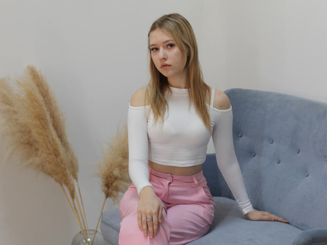 RosaVance, a blonde woman with straight hair, sits on a blue couch, wearing a white off-shoulder top and pink pants, with pampas grass to the left. Fucking tense, serious expression.