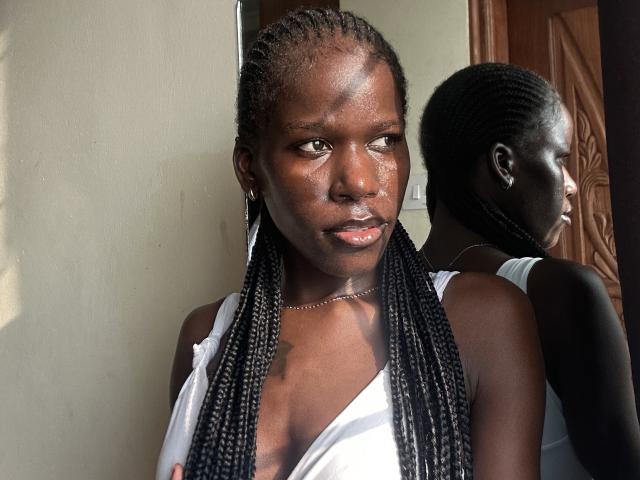 Photograph of KharlaQueen, a dark-skinned woman with braided hair, wearing a white tank top, gazing intently at the camera, reflection in the mirror showing her back, sexual tension evident.