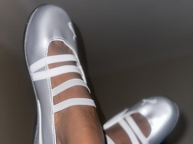 Close-up of KharlaQueen's legs showing dark brown skin and white Adidas sneakers with three white stripes. The shoes are shiny, reflecting light. The legs are slightly apart, creating tension.