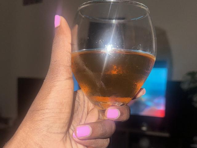 Close-up of KharlaQueen's hand holding a glass of amber liquid, with bright light reflecting off the glass. KharlaQueen's nails are painted bright pink. Focused light creates a glowing effect.