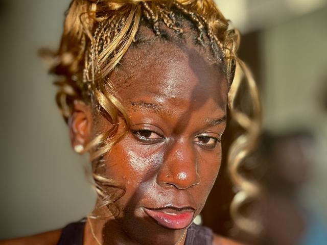 Close-up of KharlaQueen, a dark-skinned woman with golden braids, glistening skin, and pink lips. She gazes intensely, light shadows accentuating her features. Blurred background, soft focus on her face.