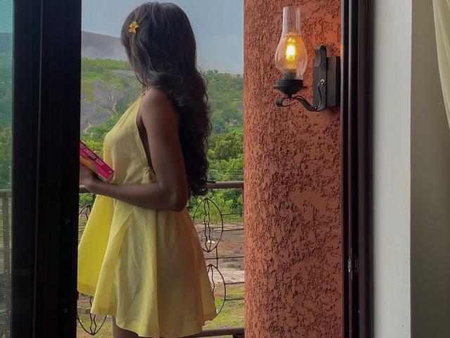 KharlaQueen está em uma porta, vestindo um vestido amarelo, segurando um objeto rosa, de costas para o observador. Cabelo comprido, flor amarela no cabelo. Lâmpada quente em uma parede texturizada. Vista externa.