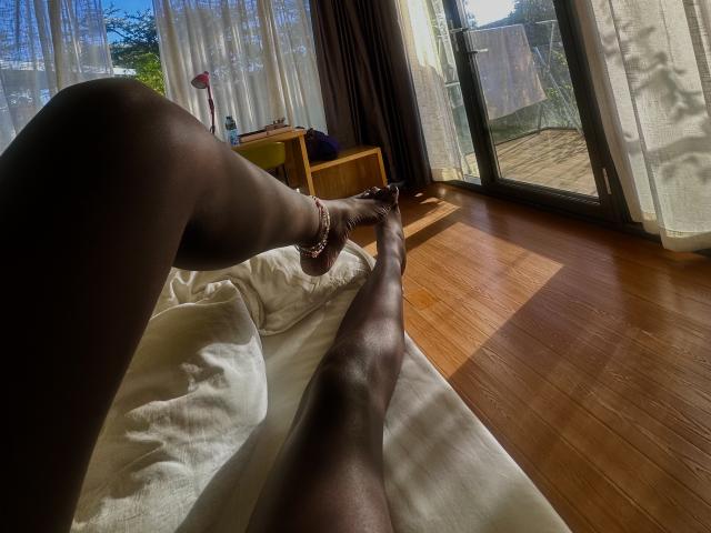 Close-up of KharlaQueen's dark-skinned legs stretched out on a white couch, sunlight filtering through sheer curtains, wooden floor, glass door, outside garden visible. Fingernails painted red, anklet on right ankle.