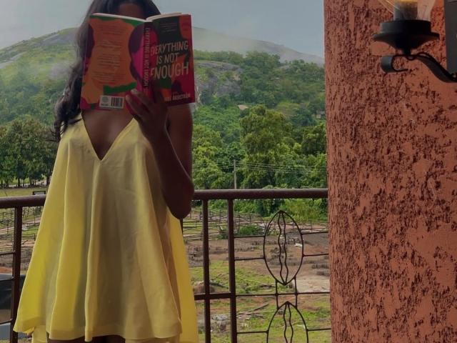 KharlaQueen, met lang donker haar, gekleed in een gele jurk met een V-hals, leest een boek met een rode omslag en staat op een balkon. Bergen en groene bomen op de achtergrond. Rechts: een textuurwand..