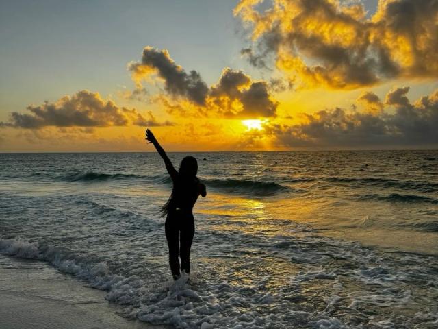 Silhouetted KharlaQueen with one arm raised, standing in shallow ocean waves at sunset. Golden clouds and orange sky contrast with dark water. Sexual tension from provocative pose, highlighted by sun's glow.