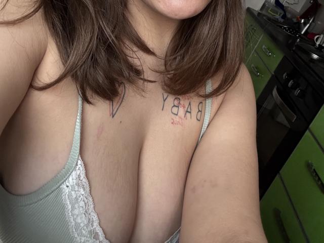 Primo piano di Kametia con capelli marrone medio, che mostra il seno con un reggiseno bianco in pizzo. Tatuaggio con la scritta "YAGA" sul petto, con lievi segni rossastri. Arredamento verde sullo sfondo. Tensione sessuale, incredibilmente eccitante..