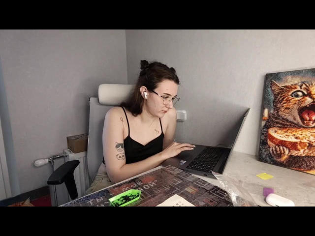 Natosha Burdine, uma mulher de pele clara, com óculos, camiseta preta e tatuagens, está sentada em uma mesa com um laptop, com um galo em seu colar, vibrador na mesa, arte colorida na parede.