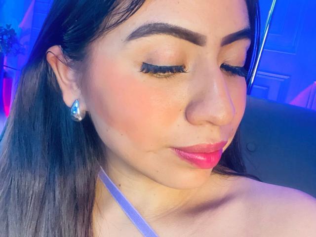 Close-up of ZoeRedds with long brown hair, closed eyes, and red lipstick. She's wearing a white strap, diamond earrings, and heavy black eyeliner. Blue and purple lights in the background. Fucking hot.