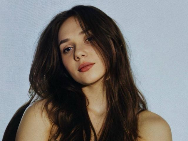 Photograph of EileneTristan, head-and-shoulders shot, dark wavy hair partially covering face, light blue background, seductive gaze, slightly parted lips, soft natural light, pale skin, subtle shadow under chin.