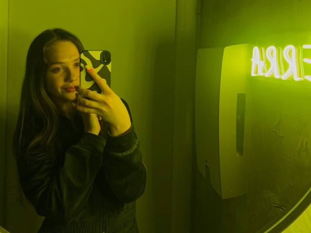 Eilene Tristan, cheveux longs et bruns, tenant un téléphone avec une lueur verte néon, portant un pull noir, reflet dans un miroir, vert. Panneau néon "NOISE", expression séduisante, éclairage jaune-vert, légère ombre sur le visage..