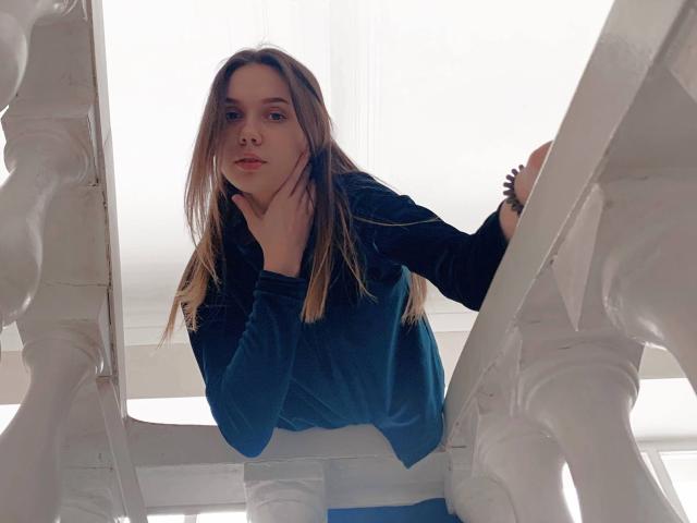 Eilene Tristan, die sich an der weißen, verzierten Balkonbrüstung abstützt, trägt ein lockeres blaues Hemd, hat hellbraunes Haar, einen intensiven Blick und eine Hand. Wangen berühren, weiches, natürliches Licht, überbelichteter Hintergrund, lässige Pose, leichte sexuelle Spannung..