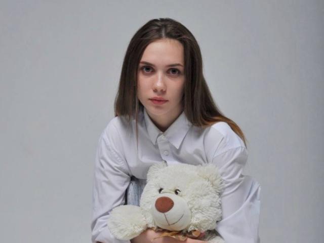 Eilene Tristan, mit langen braunen Haaren, trägt ein weißes Hemd mit Knopfleiste und hält einen flauschigen weißen Teddybären mit einer braunen Nase. mit direktem Blick in die Kamera und einem subtilen, verführerischen Ausdruck..