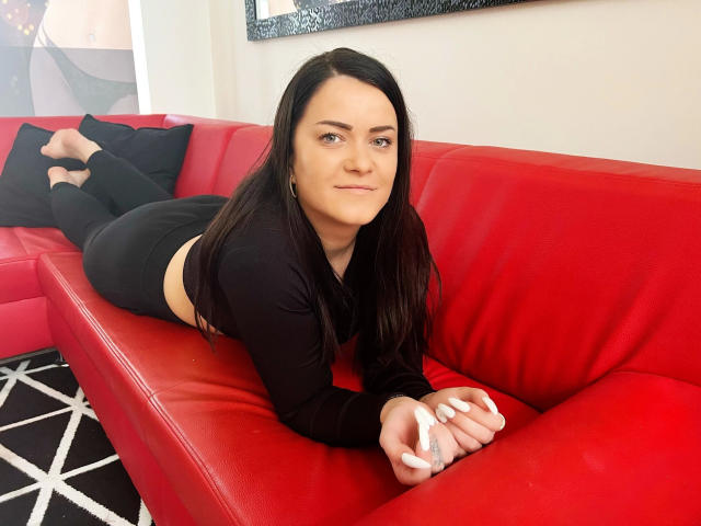 NicolSparkleX-hot lies on a red leather couch, legs bent at the knees, wearing black pants and a long-sleeve top. Her black hair cascades over her shoulders. Fucking sexy.