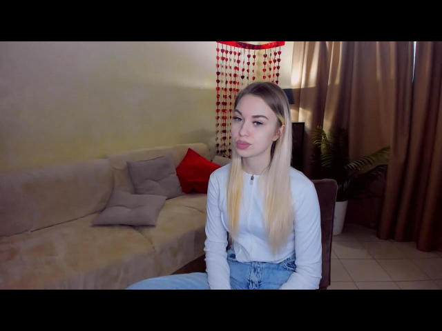 Foto van NicoleHrum zittend op een stoel, gekleed in een wit shirt met lange mouwen en blauwe jeans, met blond haar, rood rode lipstick, en een serieuze uitdrukking. De achtergrond toont een beige bank en gordijnen..