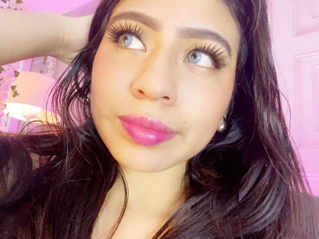 Close-up photo of ZoeRedds, looking up with wide blue eyes, glossy pink lips, and long black hair. Her hand touches her head. Bright light highlights her face. Fucking cute.